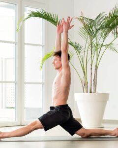 Man performing a Hanumanasna or Monkey yoga pose doing forward splits with arms raised in a high key gym with copyspace in a health and fitness concept